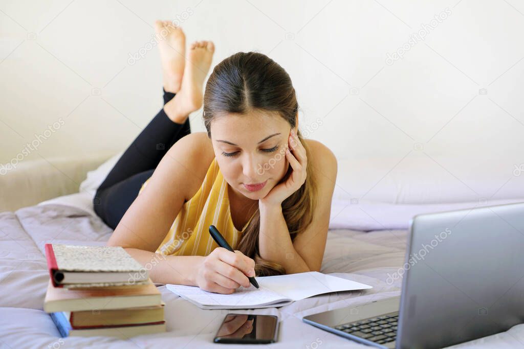 Distance learning, Homeschooling concept. Pretty teenager girl doing homework lying on bed at home. Young pretty girl writing down in notebook, studying online with laptop.