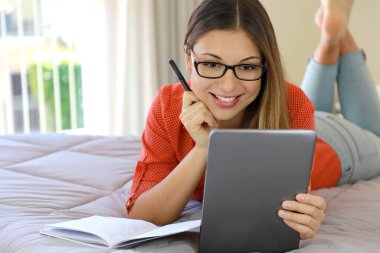 Gözlük takan Brezilyalı genç iş kadını web kamerasıyla tablet üzerinden sohbet mesafesini arıyor ya da internet kursuna katılıyor. Öğrenci kız rahat bir şekilde evde yatarak ders çalışıyor..