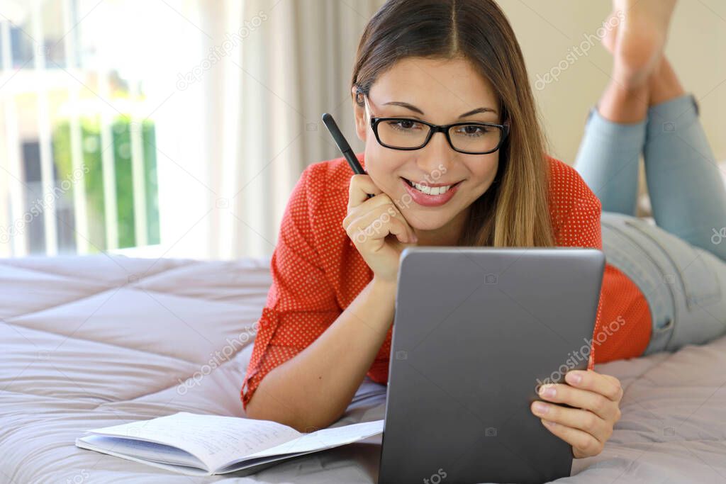 Brazilian young business woman wearing glasses video calling chat distance webinar online on tablet by webcam or attends online course. Student girl studying comfortable lying on bed from home.
