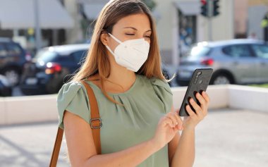 Coronavirus Hastalığı 2019 'a tepki olarak KN95 FFP2 Maskeli Genç Kadın, KOVID-19 Kişilik İzleme ve Kişilik Tanılama Programına Yardım Etmek İçin Şehir Caddesinde Akıllı Telefon Uygulaması Kullanıyor