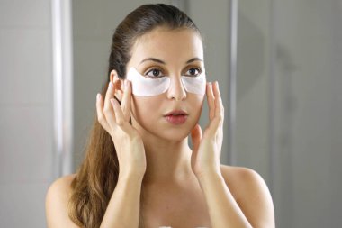 Beauty woman applying under-eye mask looking herself in the mirror in the bathroom. Skin care girl touch patches of fabric mask under eyes to reduce eye bags. clipart