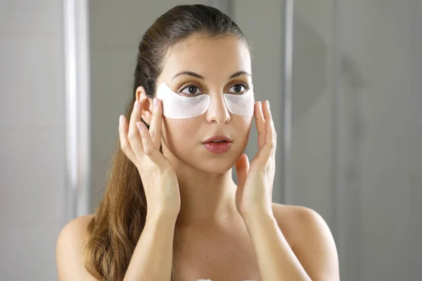 Beauty woman applying under-eye mask looking herself in the mirror in the bathroom. Skin care girl touch patches of fabric mask under eyes to reduce eye bags.