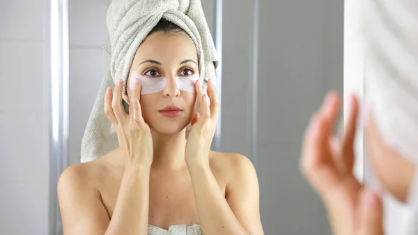 Beauty woman applying anti-fatigue under-eye mask looking herself in the mirror in bathroom. Skin care girl touch patches of fabric mask under eyes to reduce eye bags.