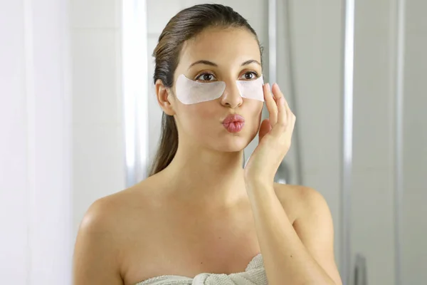 Attractive woman applying anti-fatigue under-eye mask looking and kissing herself in the mirror in the bathroom. Skin care girl touch patches of fabric mask under eyes to reduce eye bags.