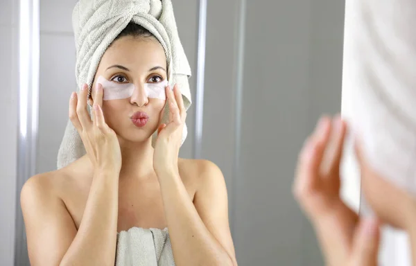 Beautiful woman applying anti-fatigue under-eye mask kissing herself in the mirror in the bathroom. Skin care girl touch patches of fabric mask under eyes to reduce eye bags.