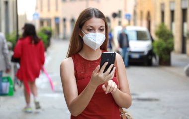 SARS-CoV-2 hastalığına karşı KN95 FFP2 maskesi takan ve Coronavirus Hastalığı 2019 'a tepki olarak Akıllı Telefon uygulaması kullanan şehir sokaklarında COVID-19 Kadın.