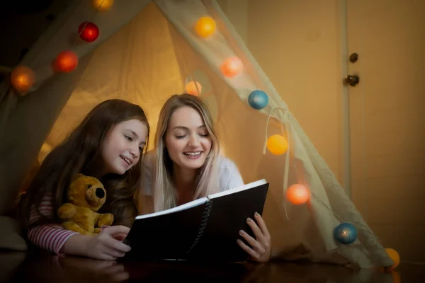 Glad liten flicka och mor ler och läser bok tillsammans i t — Stockfoto