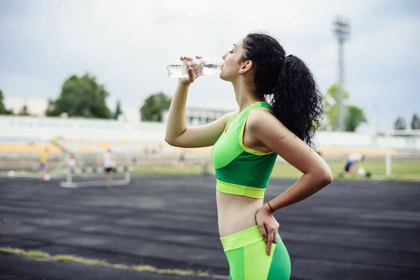 在体育场里 一头卷曲的黑发正在做运动 女孩在训练后从瓶子里喝水 — 图库照片