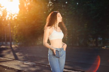 Uzun kıvırcık saçlı, beyaz bluzlu, kot pantolonlu ve telefonda konuşan bir esmerin portresi. Eğlenceli bir sohbet. Arkaplanda yeşil ağaçlar