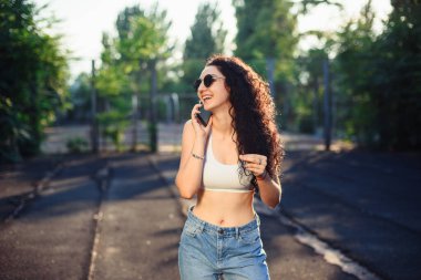 Uzun kıvırcık saçlı, gözlüklü, beyaz bluzlu ve kot pantolonlu genç bir esmer sokakta telefonla konuşuyor. Eğlenceli bir sohbet. Arkaplanda yeşil ağaçlar