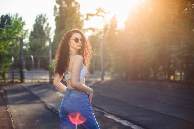 Uzun kıvırcık saçlı, gözlüklü, beyaz bluzlu ve kot pantolonlu genç bir esmer sokakta dikiliyor. Kız vurgulanıyor. Yaşam tarzı. Arkaplanda yeşil ağaçlar