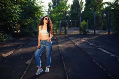 Uzun kıvırcık saçlı, gözlüklü, beyaz bluzlu ve kot pantolonlu genç bir esmer sokakta dikiliyor. Kız vurgulanıyor. Yaşam tarzı. Arkaplanda yeşil ağaçlar