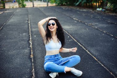 Uzun kıvırcık saçlı gözlüklü genç bir esmer, beyaz bir bluzla caddede yerde oturuyor ve elinde tablet kulaklığı olan bir kot pantolon giyiyor. Yaşam tarzı.