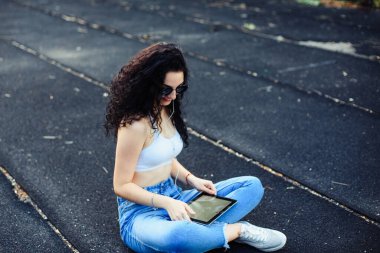 Uzun kıvırcık saçlı gözlüklü genç bir esmer, beyaz bir bluzla caddede yerde oturuyor ve elinde tablet kulaklığı olan bir kot pantolon giyiyor. Yaşam tarzı.