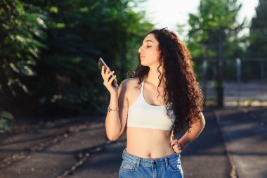 Uzun kıvırcık saçlı, beyaz bluzlu, kot pantolonlu ve telefonda konuşan bir esmerin portresi. Eğlenceli bir sohbet. Arkaplanda yeşil ağaçlar