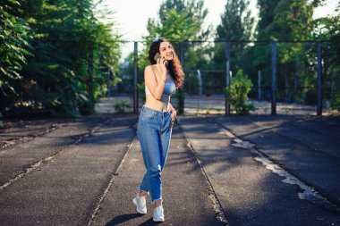 Uzun kıvırcık saçlı, beyaz bluzlu, kot pantolonlu ve telefonda konuşan bir esmerin tam boy fotoğrafı. Eğlenceli bir sohbet. Arkaplanda yeşil ağaçlar