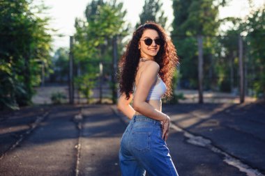 Uzun kıvırcık saçlı, gözlüklü, beyaz bluzlu ve kot pantolonlu genç bir esmer sokakta dikiliyor. Kız vurgulanıyor. Yaşam tarzı. Arkaplanda yeşil ağaçlar