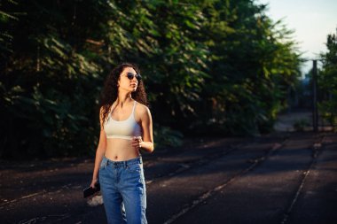 Uzun kıvırcık saçlı, gözlüklü, beyaz bluzlu ve kot pantolonlu genç bir esmer sokakta dikiliyor. Kız vurgulanıyor. Yaşam tarzı. Arkaplanda yeşil ağaçlar