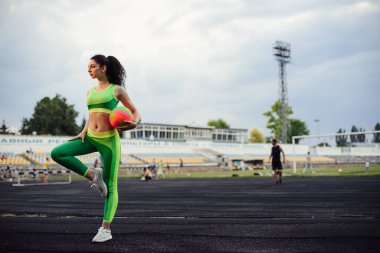 Stadyumda kaslarını esneten güzel kıvırcık kız. Topla ısınmaya başla. Kız spora gidiyor. Açık yeşil eşofman. Yatay fotoğraf.