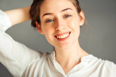 Tatmin olmuş gülümseyen bir kız ellerini gri bir arka planda tutuyor, yakın plan. Çıplak makyaj, rötuş yapmadan..