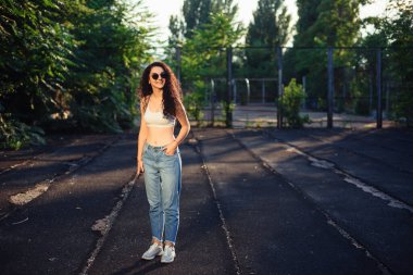 Uzun kıvırcık saçlı, gözlüklü, beyaz bluzlu ve kot pantolonlu genç bir esmer sokakta dikiliyor. Kız vurgulanıyor. Yaşam tarzı. Arkaplanda yeşil ağaçlar