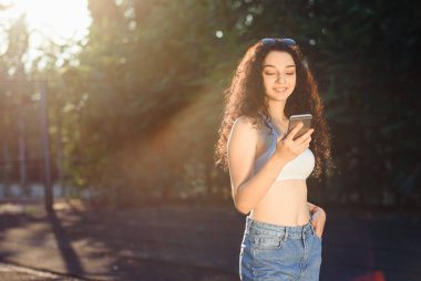 Uzun kıvırcık saçlı, beyaz bluzlu, kot pantolonlu ve telefonda konuşan bir esmerin portresi. Eğlenceli bir sohbet. Arkaplanda yeşil ağaçlar