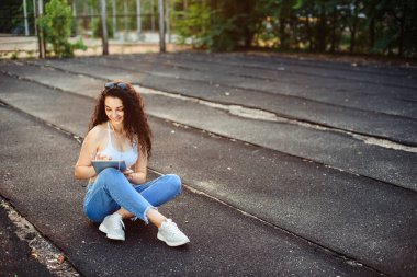 Uzun kıvırcık saçlı gözlüklü genç bir esmer, beyaz bir bluzla caddede yerde oturuyor ve elinde tablet kulaklığı olan bir kot pantolon giyiyor. Yaşam tarzı.