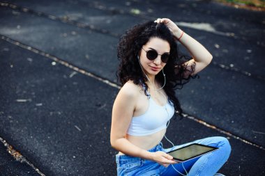 Uzun kıvırcık saçlı gözlüklü genç bir esmer, beyaz bir bluzla caddede yerde oturuyor ve elinde tablet kulaklığı olan bir kot pantolon giyiyor. Yaşam tarzı.