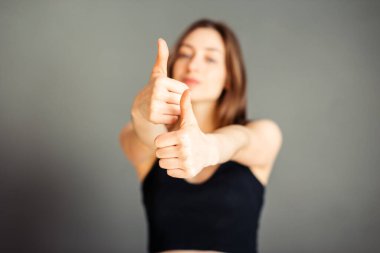 Saçları siyah bluzlu kız başparmağını kaldırıyor. Gri arka planda mutlu. Makyajsız, rötuşsuz.