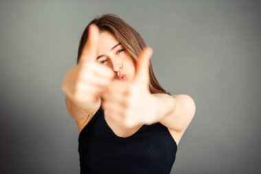 Saçları siyah bluzlu kız başparmağını kaldırıyor. Gri arka planda mutlu. Makyajsız, rötuşsuz.