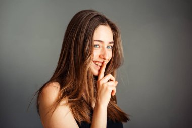 A girl in a black top with her hair is asking for peace and quiet, gesturing with a finger in front of her mouth, saying shhh with a smile on her face on a gray background. clipart