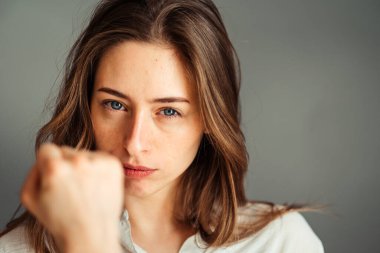 Ciddi kız yumruğunu gösteriyor. Yüze yakın çekim. Gri bir arka planda. Gevşek doğal saçlar. Makyajsız, rötuşsuz.