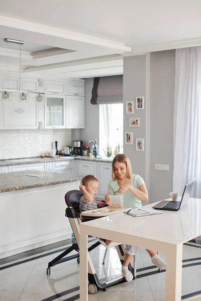 Portrait of mid age caucasian blond woman with child being busy with computer and feeding the baby. They are at home, wearing pyjamas