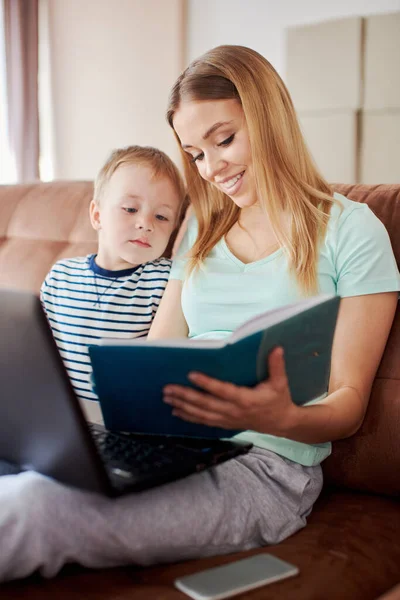 Educational and entertainment laptop app concept. Mother and son are using laptop, and making drawings in notepad. They are cosy sitting at the sofa at home