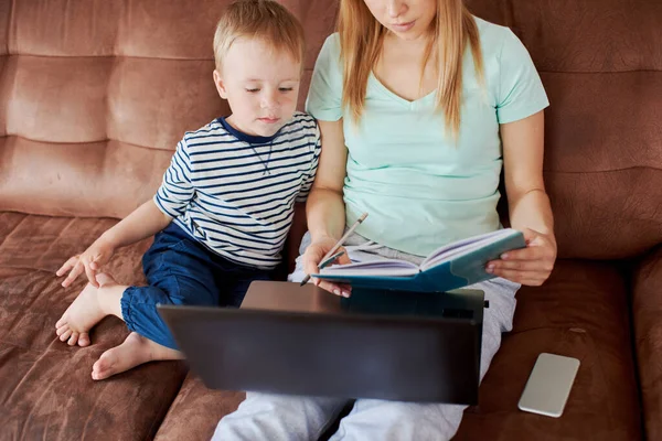Educational and entertainment laptop app concept. Mother and son are using laptop, and making drawings in notepad. They are cosy sitting at the sofa at home
