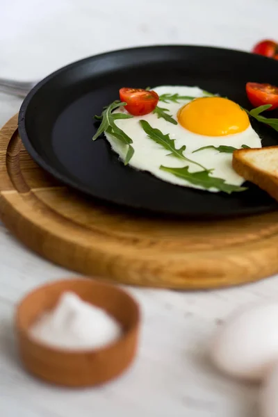 Жареное куриное яйцо в черной сковороде с помидорами черри, рукколой, солью. Завтрак на белом деревянном столе . — стоковое фото