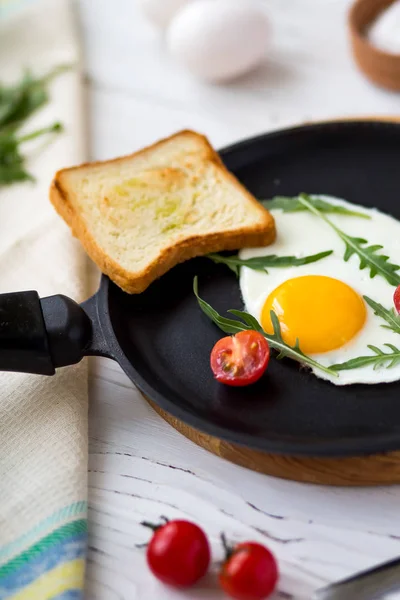 Жареное куриное яйцо в черной сковороде с помидорами черри, рукколой, солью. Завтрак на белом деревянном столе . — стоковое фото