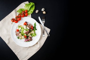 Biftek, peynir, ot ve bıldırcın yumurtası beyaz tabakta, çatal ve bıçağın yanında. Siyah masanın üzerinde. Çeri domates ve sebzeli bıldırcın yumurtasıyla süslenmiş. Yukarıdan manzara. Boşluğu kopyala