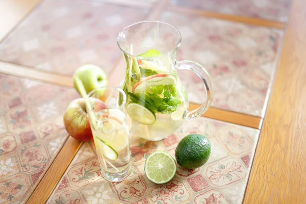 Lemonade on the table in a clear jug and glass. With pieces of fruit. Apples and limes on a ceramic tile table. Natural daylight. Close up
