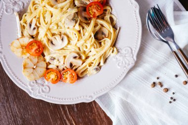 Mantarlı, domatesli ve sarımsaklı kremalı İtalyan makarnası. Kahverengi bir sehpanın üstündeki çatal bıçak takımının içinde peçete ve baharatlarla. Üst görünüm