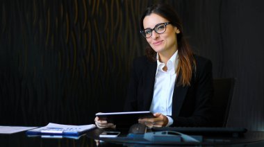 Genç düşünceli sekreter masasında dijital tablet kullanarak gülümsüyor.