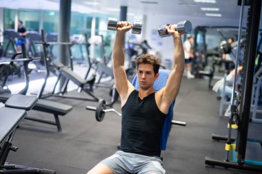 Vücut geliştirici, spor salonunda kollarını ve omuzlarını çalıştırmak için dambılları kaldırıyor.