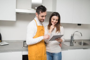 Mutfakta pişirmek için tablette yemek tarifi arayan mutlu bir çift.