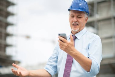 Sinirli ve stresli mimar inşaat alanının önünde cep telefonuna bakıyor.