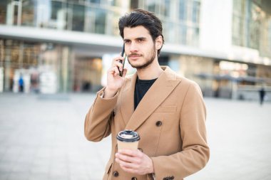 Adam telefonda konuşuyor ve şehir meydanında yürürken elinde bir fincan kahve tutuyor.