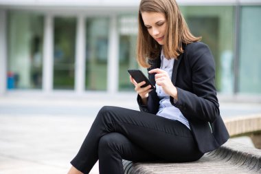 İş kadını ofisinin önündeki bankta otururken cep telefonunu kullanıyor.