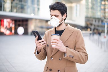 Maskeli genç adam cep telefonunu kullanıyor ve bir şehirde yürürken bir fincan kahve tutuyor, koronavirüs ve salgın hastalık konsepti.