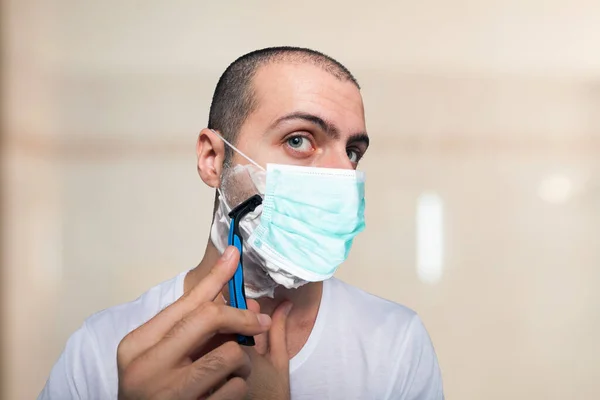 マスクをしながら髭を剃る使い捨てカミソリを使う男コロナウイルスパンデミックの概念 — ストック写真