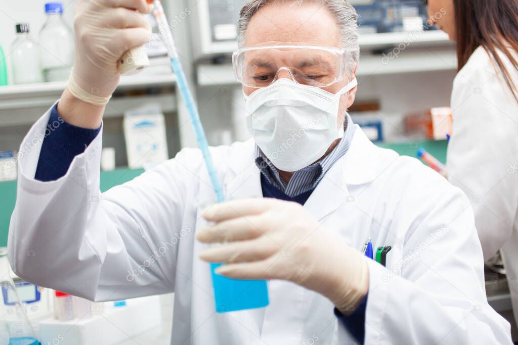 Close-up portrait of a male researcher holding flask with blue liquid in the lab, coronavirus research concept