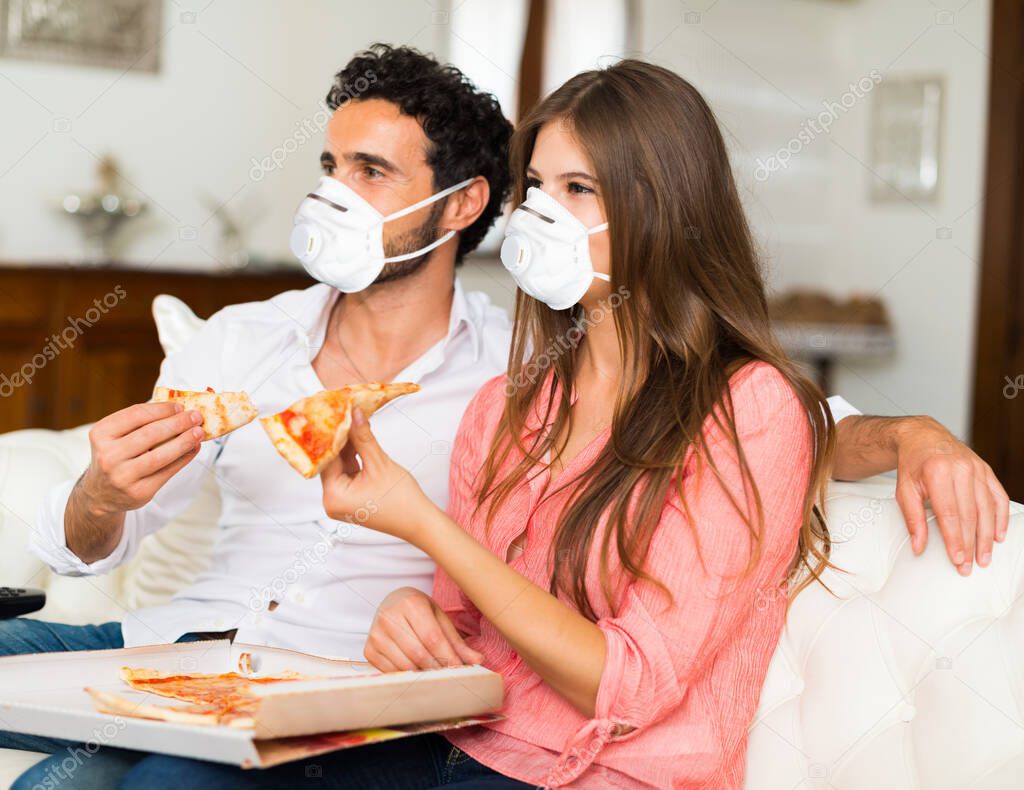 Couple eating pizza at home while wearing a mask, coronavirus concept
